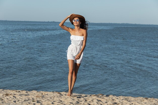 Hermosa chica de pelo oscuro con sombrero de vestir blanco y gafas de sol caminando por la playa de arena cerca del mar en un día soleado