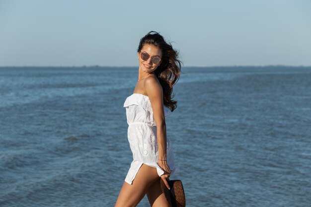 Hermosa chica de pelo oscuro con vestido blanco y gafas de sol sostiene el sombrero en la mano y camina cerca del mar en un día soleado. .