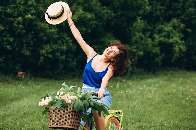 Hermosa chica de pelo rizado en un sombrero sonríe y sostiene bicicleta con cesta de flores