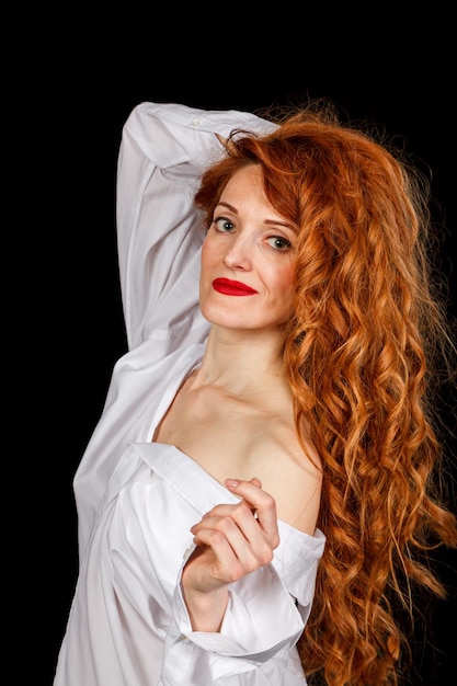 Hermosa chica con el pelo rojo en una camisa blanca y un sombrero sobre un fondo negro