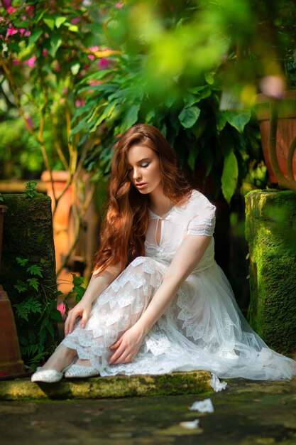 Foto hermosa chica de pelo rojo con un vestido de encaje blanco de pie en un jardín con flores de colores.