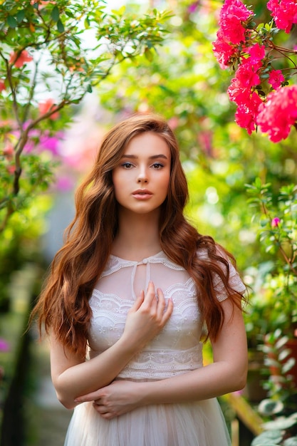 Hermosa chica de pelo rojo con un vestido de encaje blanco de pie en un jardín con flores de colores.