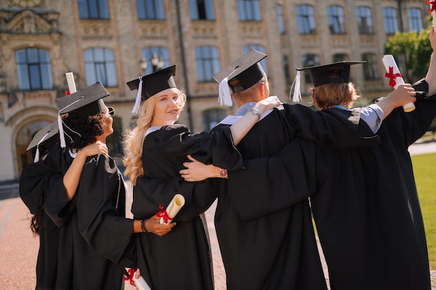 Hermosa chica de pie con sus amigos frente a su universidad después de la ceremonia de graduación