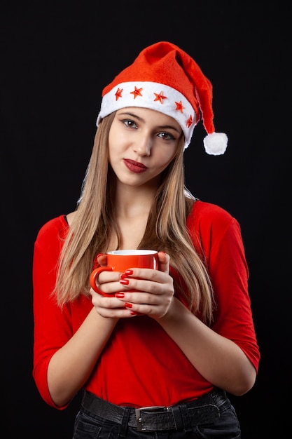 Hermosa chica posando con bebida caliente en taza roja sobre fondo negro en traje de año nuevo. Celebracion.