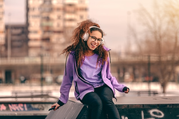 Hermosa chica de raza mixta adolescente inconformista con gran sonrisa toothy escuchando música y sentado en el skate park.