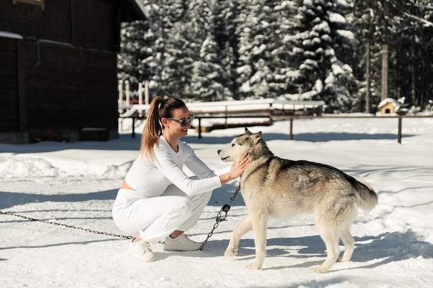 Una hermosa chica con ropa blanca y gafas de sol se divierte jugando con un husky esponjoso y alegre en el fondo de la naturaleza en un perro de invierno con correa