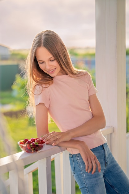 Hermosa chica, rubia, adolescente con cabello largo en una noche de verano come fresas en la terraza