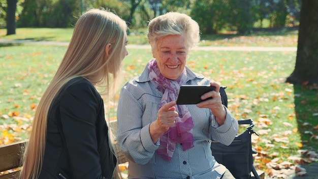 Hermosa chica rubia con anciana sentada en el banco y mirando algo en el teléfono inteligente