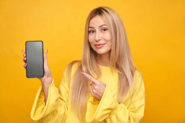 Hermosa chica rubia apuntando a la pantalla del teléfono en blanco y sonriendo aislado sobre fondo amarillo de estudio