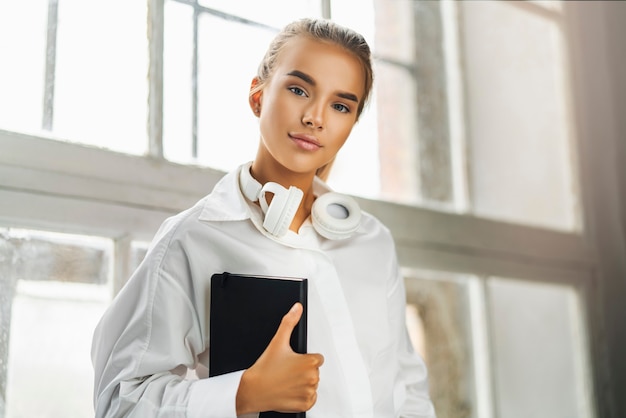 Foto hermosa chica rubia en camisa blanca con auriculares con cuaderno negro.