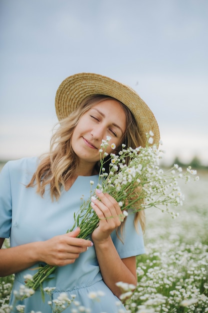 Hermosa chica rubia en un campo de margaritas. Mujer con un vestido azul en un campo de flores blancas. Niña con un ramo de margaritas. Foto tierna de verano en el pueblo. flores silvestres niña en un sombrero de paja