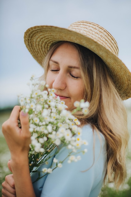 Hermosa chica rubia en un campo de margaritas. Mujer con un vestido azul en un campo de flores blancas. Niña con un ramo de margaritas. Foto tierna de verano en el pueblo. flores silvestres niña en un sombrero de paja