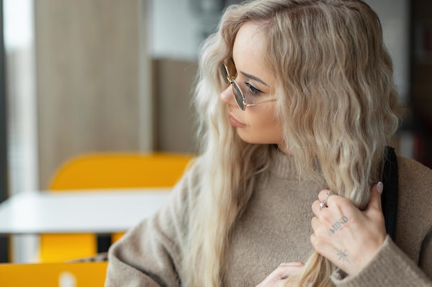 Hermosa chica rubia elegante con gafas de sol vintage en un suéter beige de moda se sienta en un café y se peina