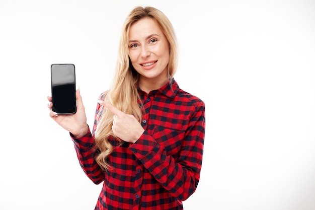 Hermosa chica rubia emocionada en puntos casuales en la pantalla del teléfono inteligente y sonrisas aisladas sobre fondo blanco de estudio