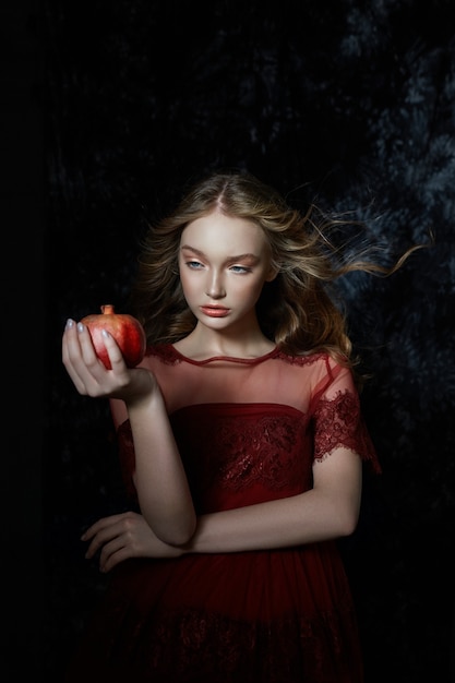 Hermosa chica rubia con fruta de granada en sus manos. Primavera retrato de una niña con un vestido rojo rompiendo una granada, jugo que fluye por sus manos