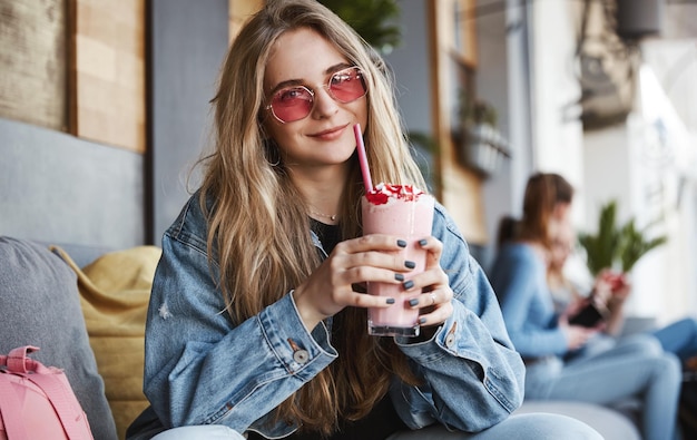 Hermosa chica rubia en gafas de sol bebiendo batido en café ter