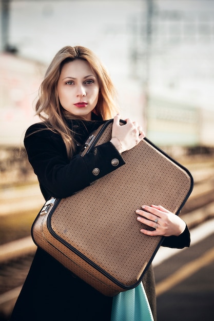 Hermosa chica rubia con una maleta esperando un tren en la estación. mirada romántica y delicada