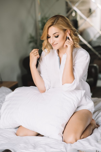 Hermosa chica rubia modelo sentada en una cama blanca con una almohada en una camisa escucha disfrutando de la música a través de auriculares.