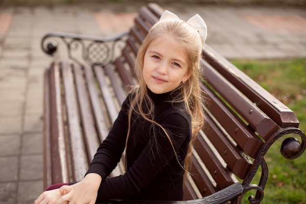 una hermosa chica rubia con el pelo largo y un vestido negro está sentada en un banco del parque