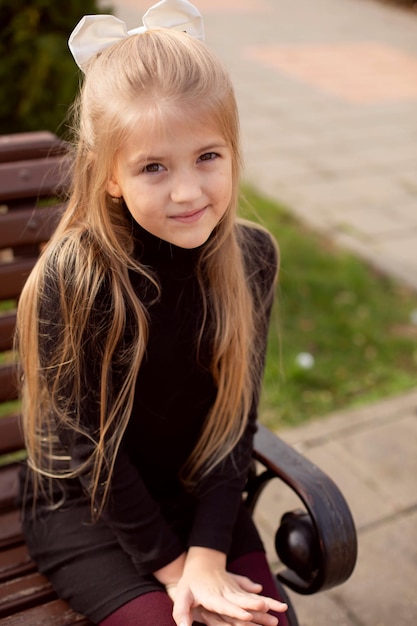 una hermosa chica rubia con el pelo largo y un vestido negro está sentada en un banco del parque