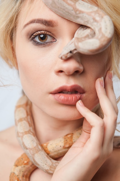 Hermosa chica rubia y la serpiente Boa constrictoras alrededor de su rostro sobre fondo gris