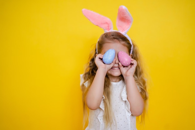 Hermosa chica rubia sonriente con orejas de conejo sosteniendo un huevo de Pascua en sus manos cierra los ojos en un niño de fondo amarillo celebra Pascua Foto de alta calidad