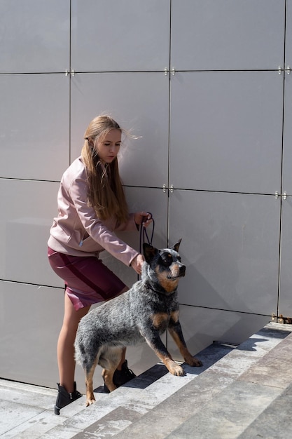 una hermosa chica rubia se para y sostiene a un perro sanador con una correa en la ciudad