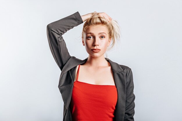 Una hermosa chica rubia sostiene su cabello y hace su propio peinado con la emoción de la sorpresa. Mujer en chaqueta negra y top rojo sobre fondo gris aislado para publicidad. Piercing de naríz