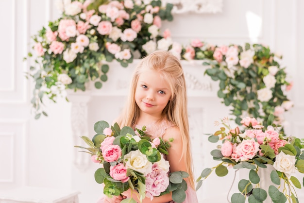 Hermosa chica rubia en un suave vestido púrpura sobre un fondo de flores posando. Modelo de bebé lindo en la imagen de una princesa. Lleva un vestido lila completo