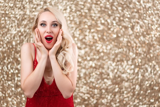 Hermosa chica rubia en un vestido de noche rojo posando Sorprendido sosteniendo sus manos cerca de su cara Fondo dorado brillante festivo