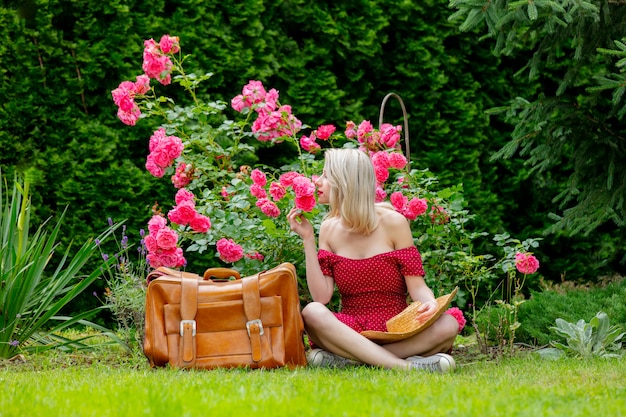 Hermosa chica rubia en vestido rojo con maleta en un jardín.