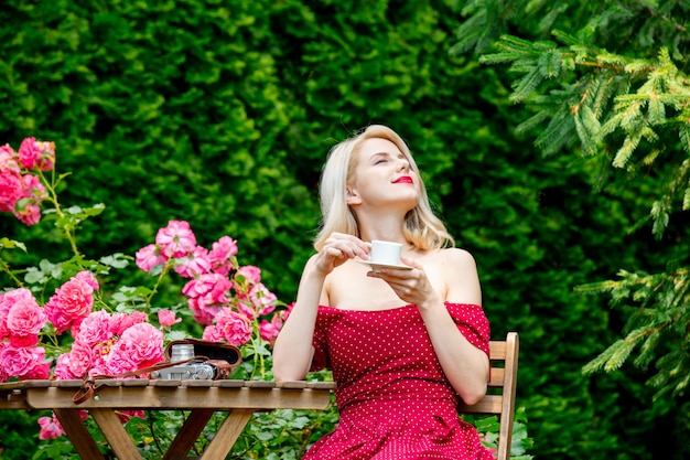 Hermosa chica rubia en vestido rojo tomando un café en un jardín.