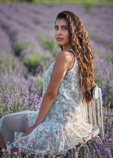 Hermosa chica en un sarafan blanco se sienta en una silla de madera y posa en medio de un campo de lavanda.