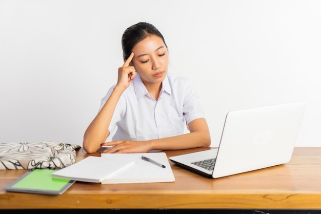 Hermosa chica de secundaria sentada en el escritorio en pose de pensamiento mientras usa la computadora portátil en el fondo blanco