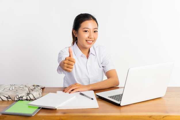 Hermosa chica de secundaria sentada en el escritorio con los pulgares hacia arriba mientras usa la computadora portátil en el fondo blanco
