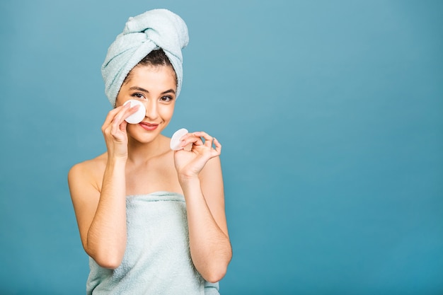 Hermosa chica sensual limpiando su rostro con un hisopo de algodón. Foto de niña después del baño en bata de baño y toalla en la cabeza aislada sobre fondo azul.