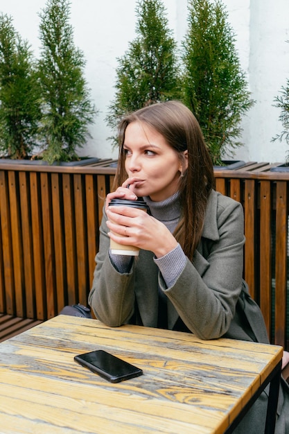 Hermosa chica sentada en la cafetería con teléfono inteligente en la mesa sosteniendo café en las manos y bebiendo