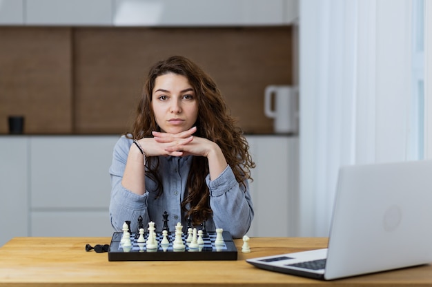 Hermosa chica sentada en casa y jugando al ajedrez en línea con una computadora portátil