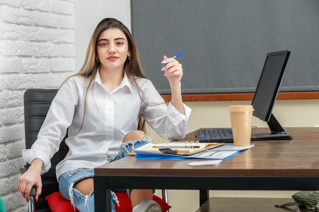 Una hermosa chica sentada detrás del escritorio y sosteniendo un bolígrafo mientras mira a la cámara