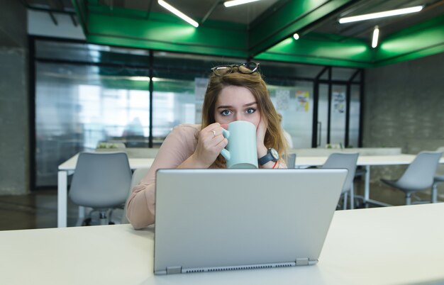 Hermosa chica sentada en el escritorio en la oficina y tomando café de la taza mientras trabajaba en una computadora.