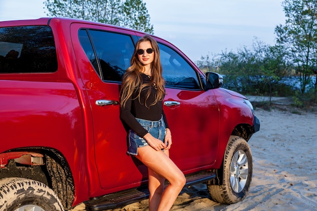 Hermosa chica sexy con el pelo largo en pantalones cortos de mezclilla cortos y un traje negro posando cerca de una camioneta roja. Chica posando cerca del coche