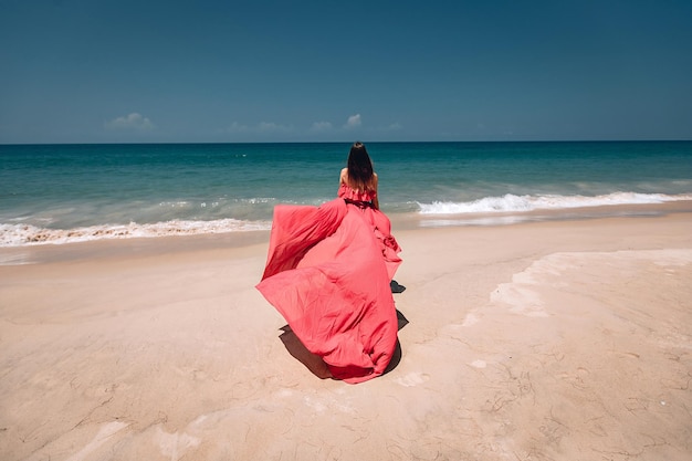 Hermosa chica en la soleada playa exótica junto al océano. La joven luce un increíble vestido rojo ondeando en la ligera brisa, vista trasera; concepto de moda.