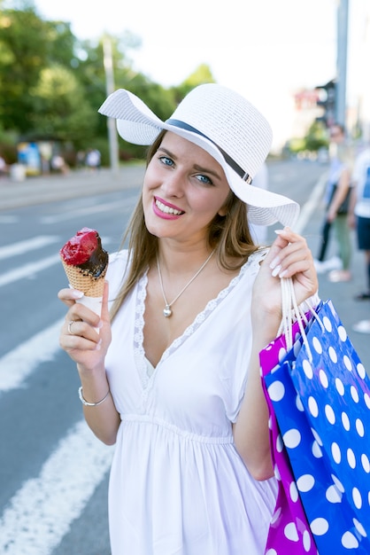 Hermosa chica con sombrero blanco y bolsas de compras comiendo un helado en la calle de la ciudad