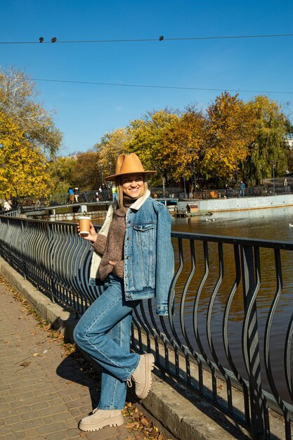 Una hermosa chica con un sombrero camina en un parque de otoño con café