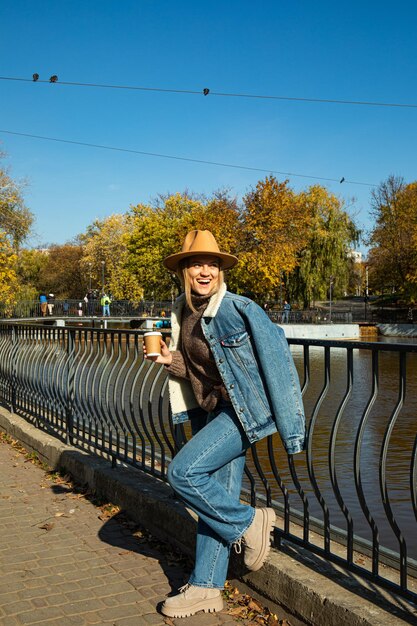 Una hermosa chica con un sombrero camina en un parque de otoño con café