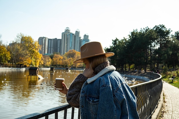 Una hermosa chica con sombrero camina en un parque de otoño con café