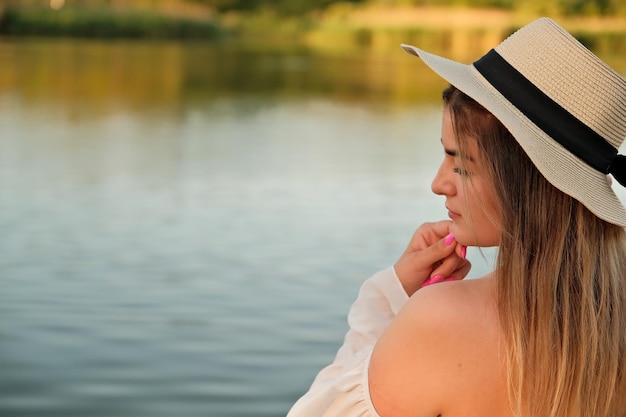 Una hermosa chica con un sombrero se encuentra en la orilla con el sombrero en la mano.