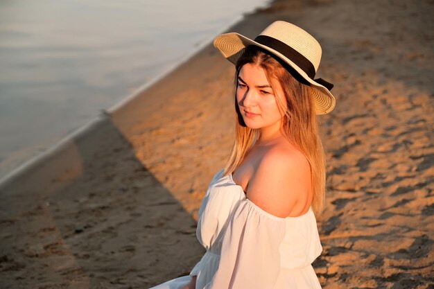 Una hermosa chica con un sombrero se encuentra en la orilla con el sombrero en la mano.