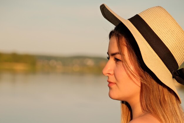 Una hermosa chica con un sombrero se encuentra en la orilla con el sombrero en la mano.