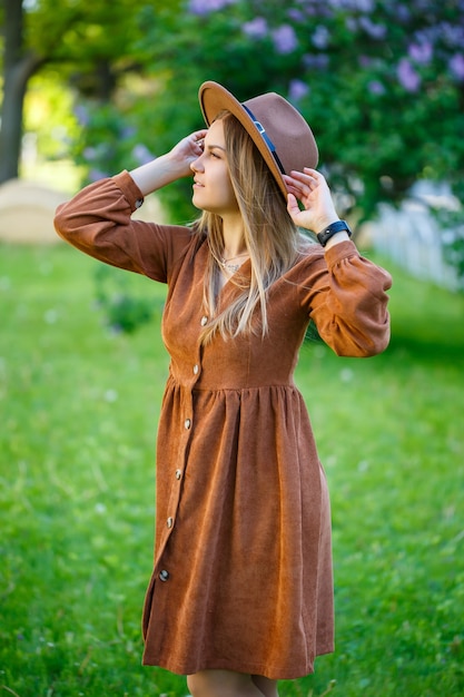 Hermosa chica con un sombrero marrón de pie cerca de un arbusto lila. Mujer joven cerca de un árbol floreciente.